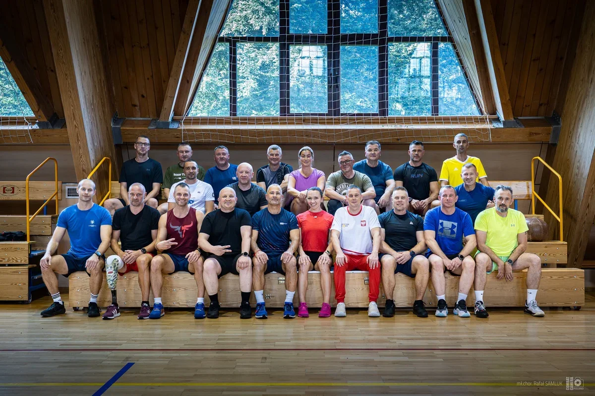 Grupa osób w strojach sportowych pozuje do pamiątkowego zdjęcia w hali sportowej z drewnianą konstrukcją dachu i dużym oknem.