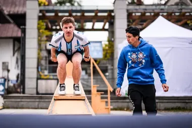 Mężczyzna w koszulce sportowej przygotowuje się do skoku na drewnianej rampie, a obok stoi mężczyzna w niebieskiej bluzie z kapturem.